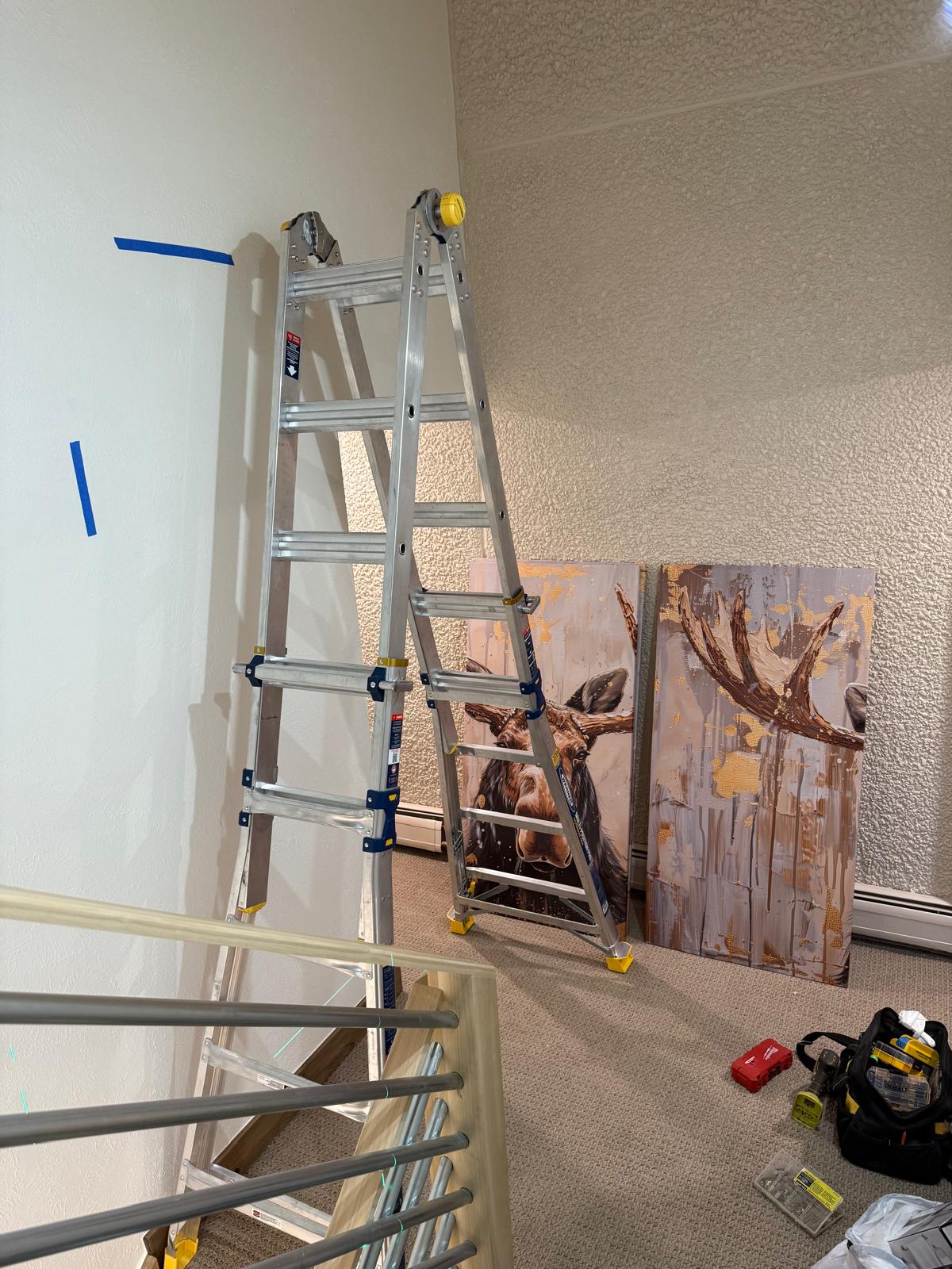 Framed canvas panels staged against a stairwell wall with the ladder set for hanging — picture hanging service in Denver