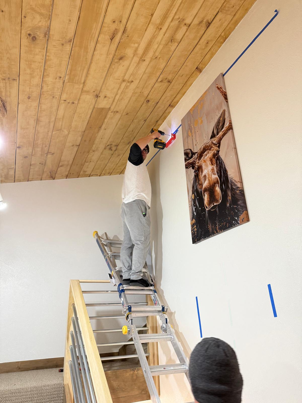 Technician on a ladder aligning a large canvas print on a high stairwell wall — picture hanging service in Denver